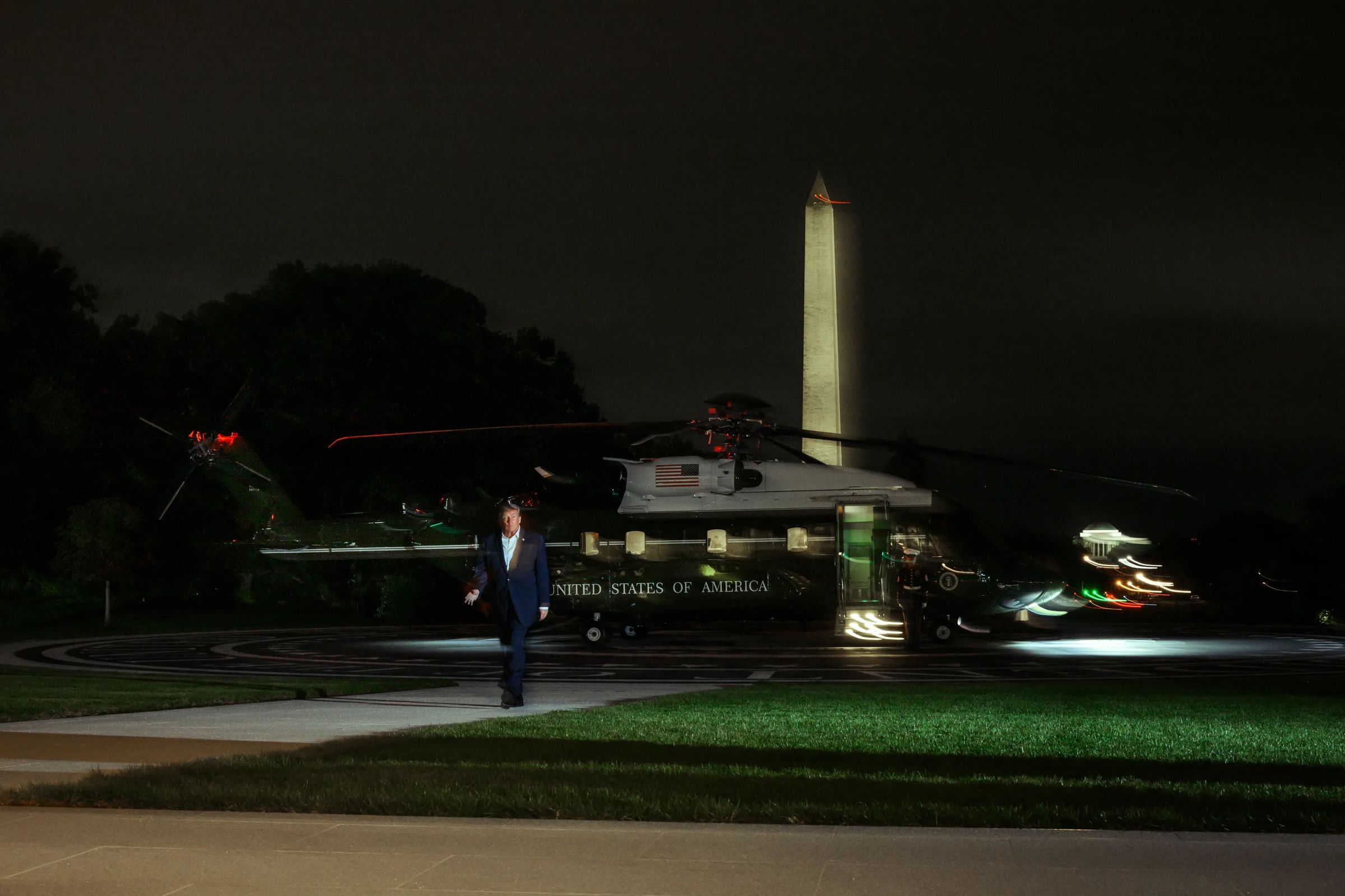 President Trump walks across the South Lawn at night after stepping off Marine One, with the Washington Monument lit up behind him.